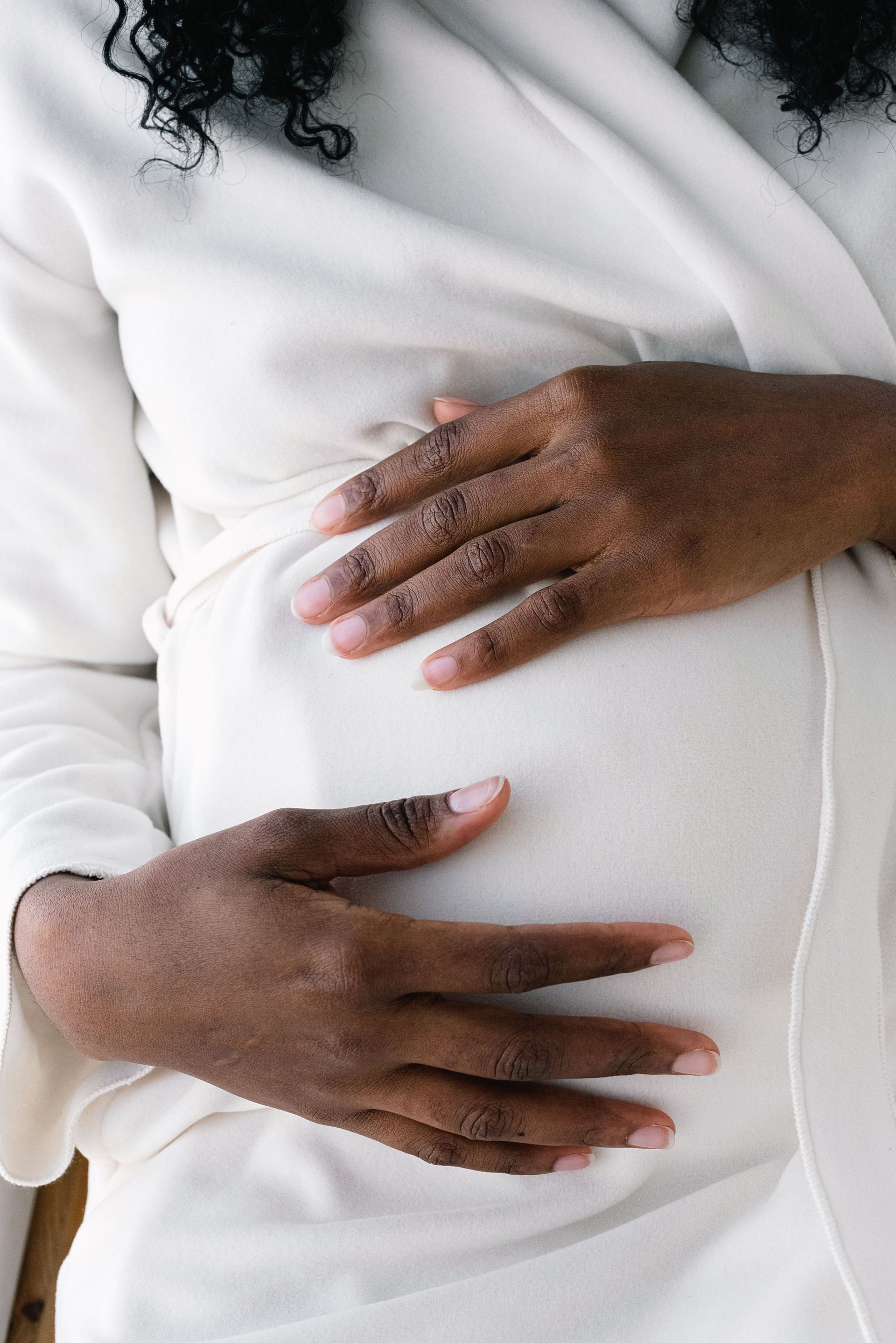 Unrecognizable African American pregnant female with dark hair in white clothes touching tummy gently with hands while standing in light room